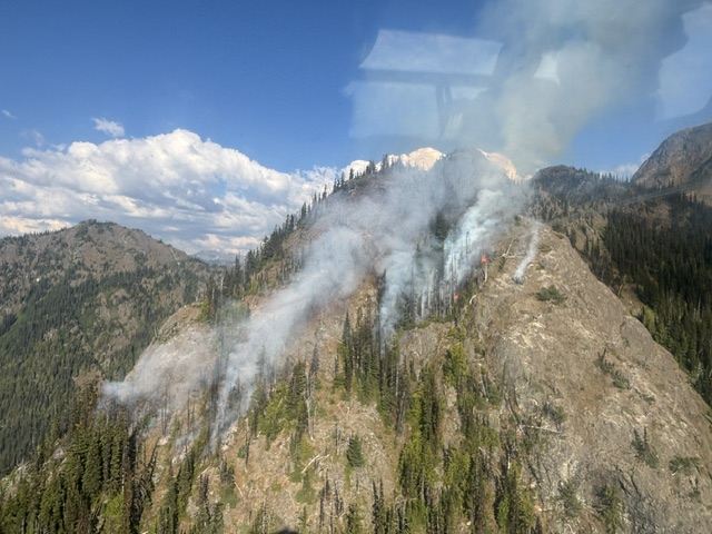 A mountain with several plumes of smoke and sparse trees under partly cloudy sky.
