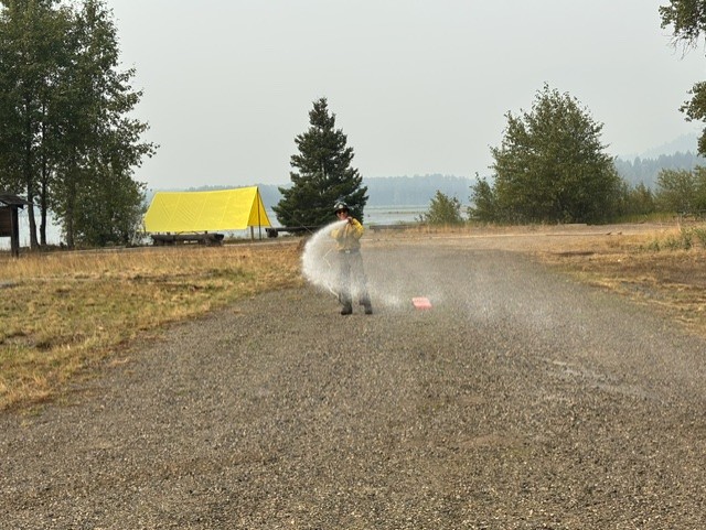Firefighter on a gravel path sprays water from a hose near a yellow tent and trees by a lake.