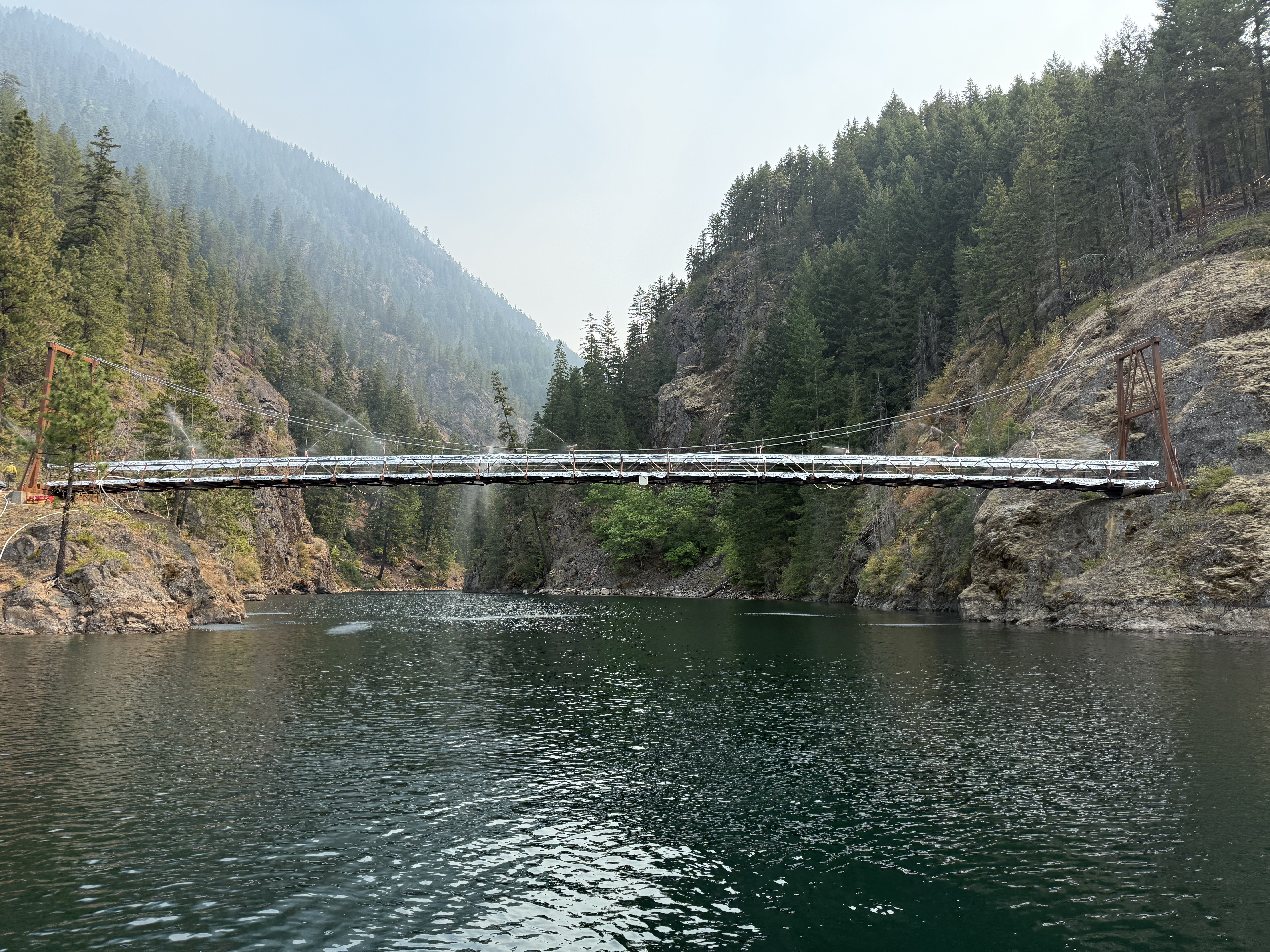 Suspension bridge in structure protection wrapping spanning a narrow lake surrounded by forested mountains under a hazy sky.
