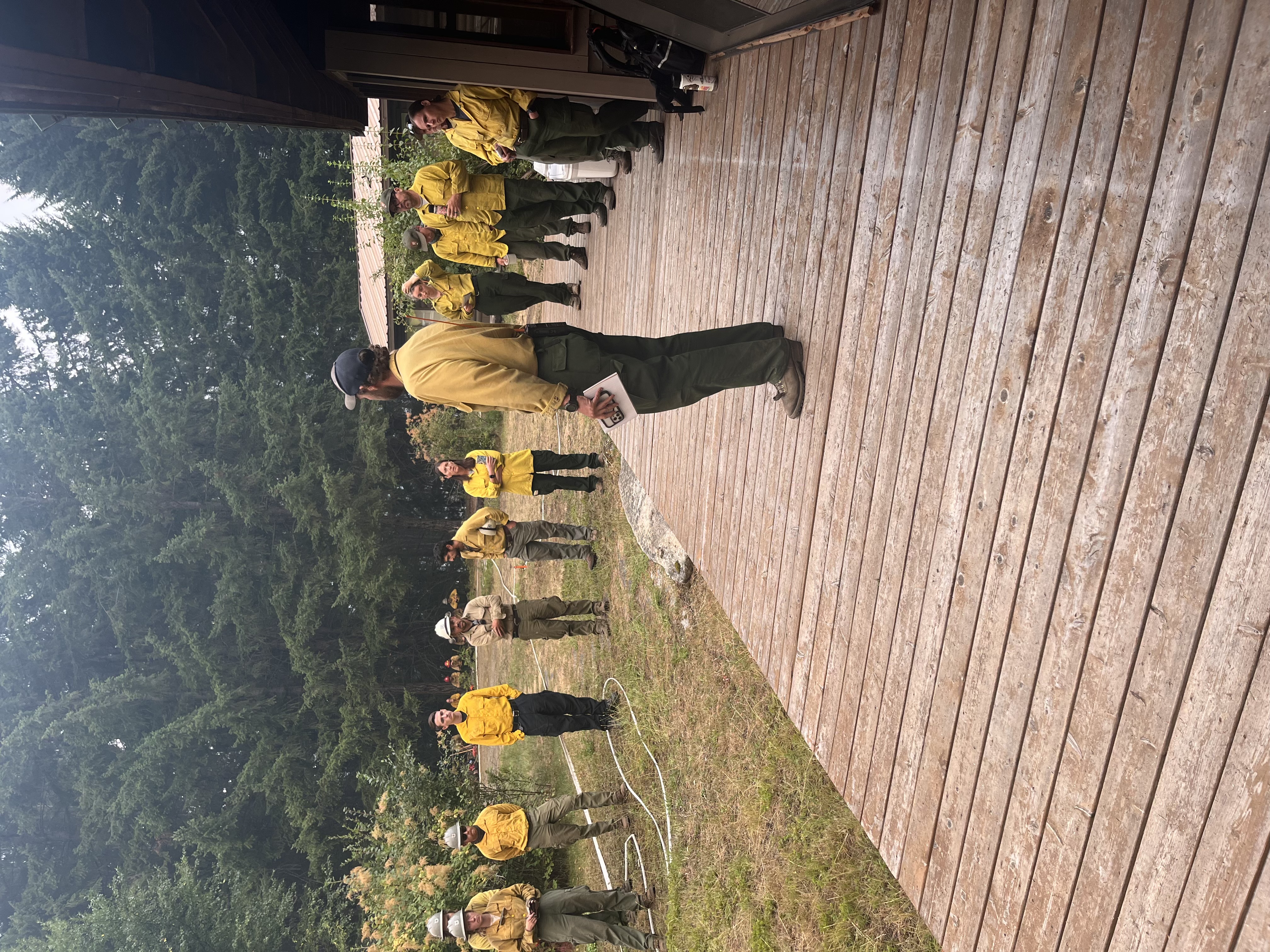 A group of people in yellow shirts and green pants stand around a building looking at the person on the wooden deck.