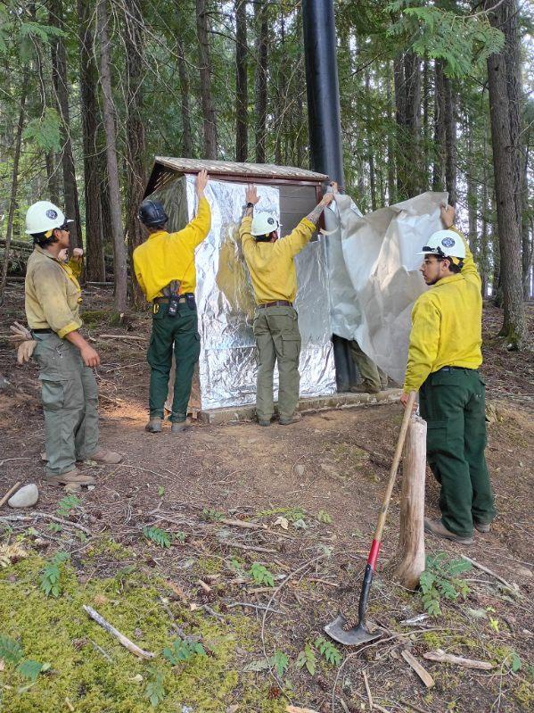 Firefighters in yellow shirts wrap a building in structural fire protection in a forested area.