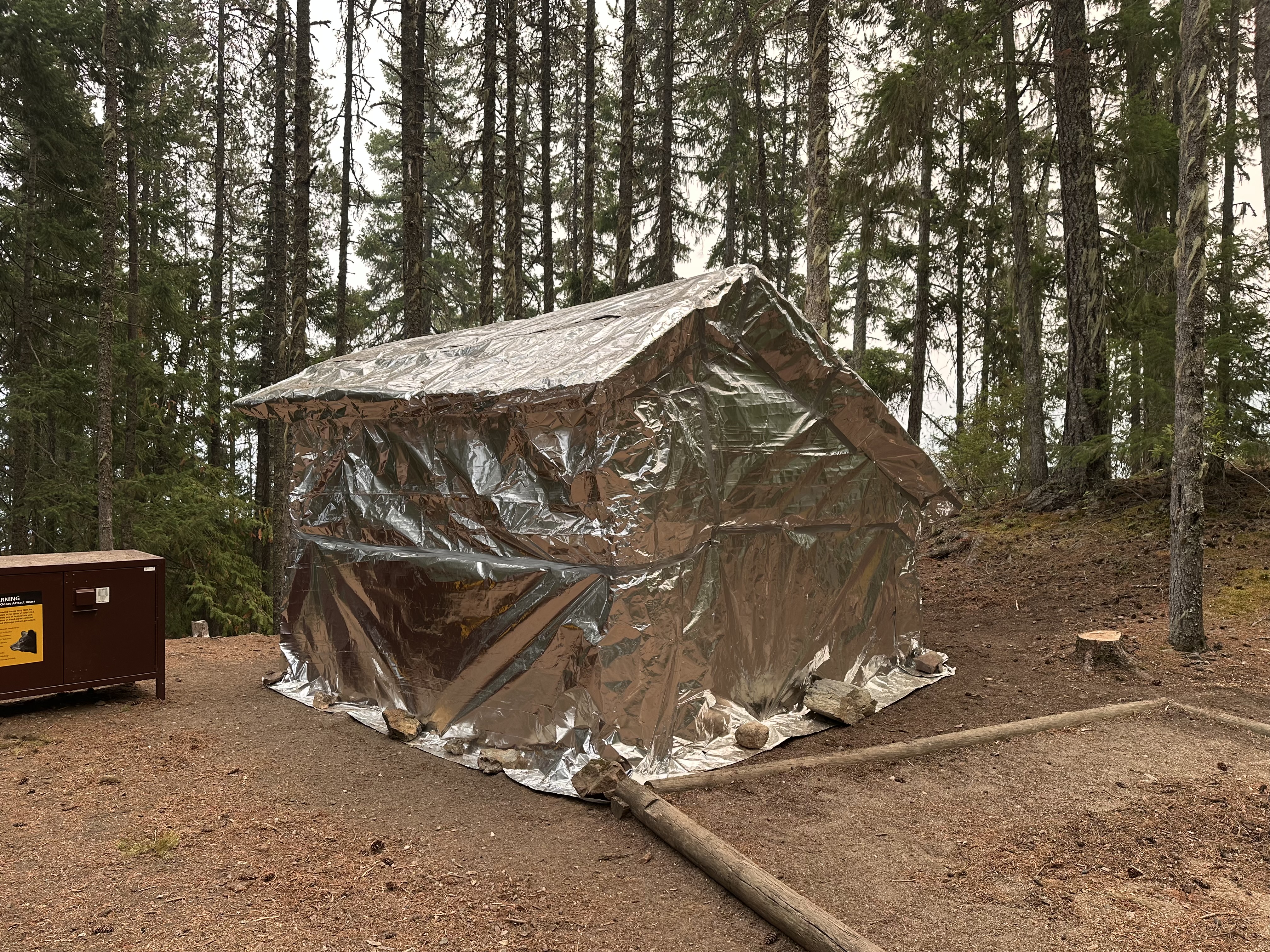 Structure wrapped in shiny aluminum material with tent pad in front and a brown bear box with sign to the left.