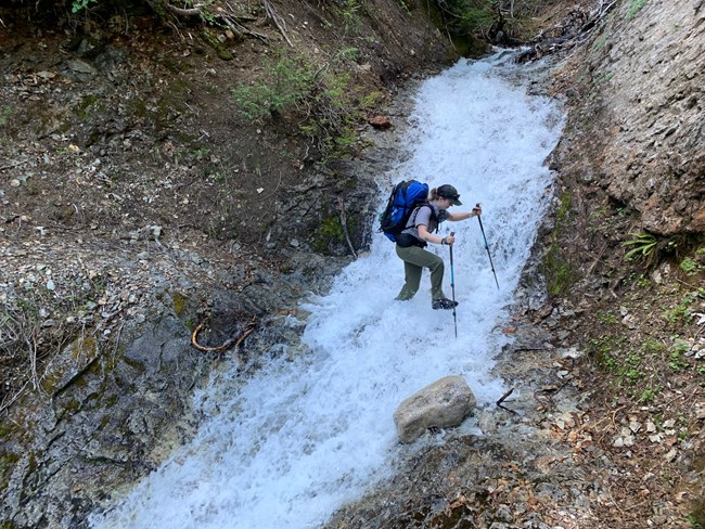 A person fords a swift creek along a trail