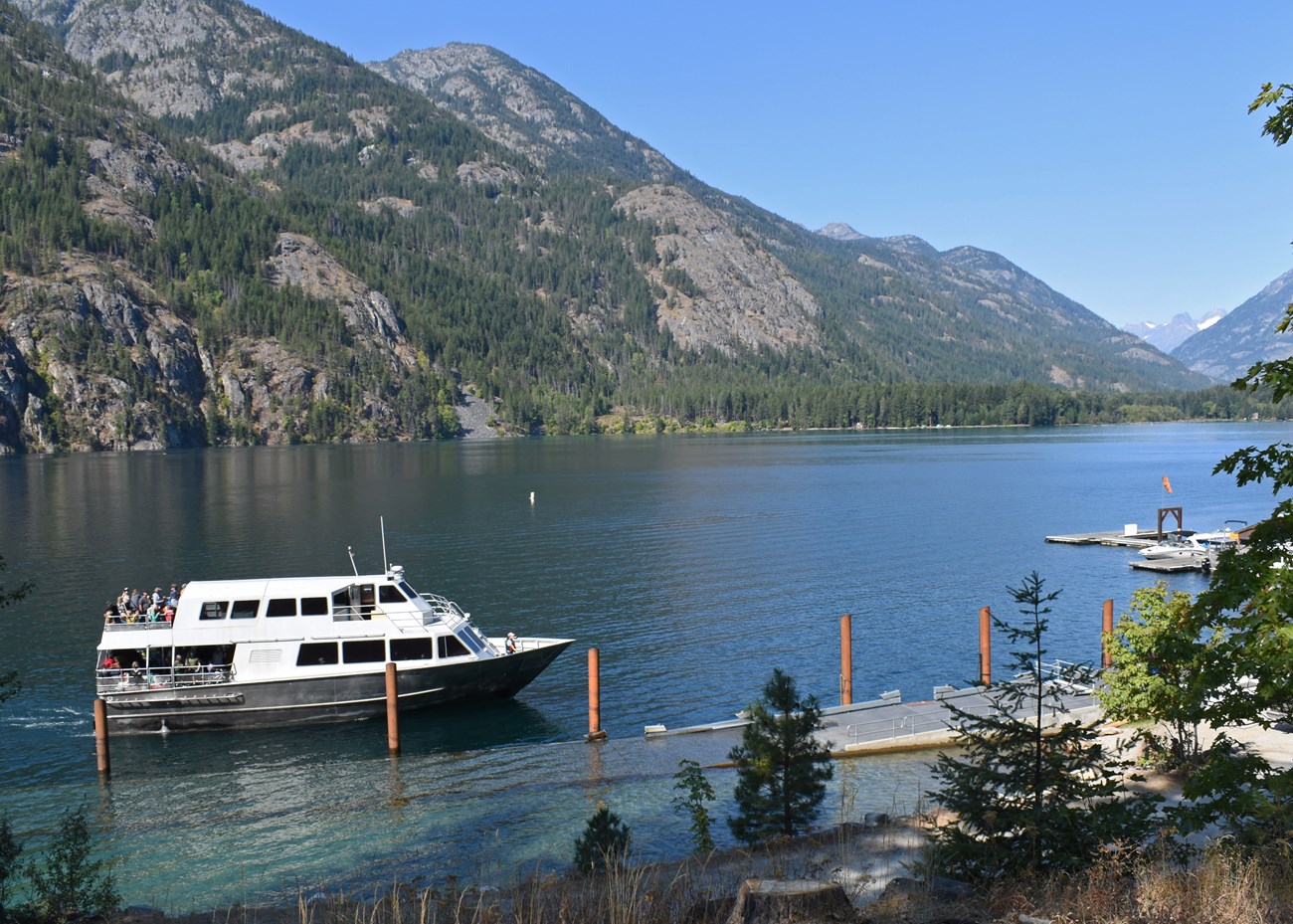 A silver ferry arrives at a dock on a large lake with mountains in the distance.