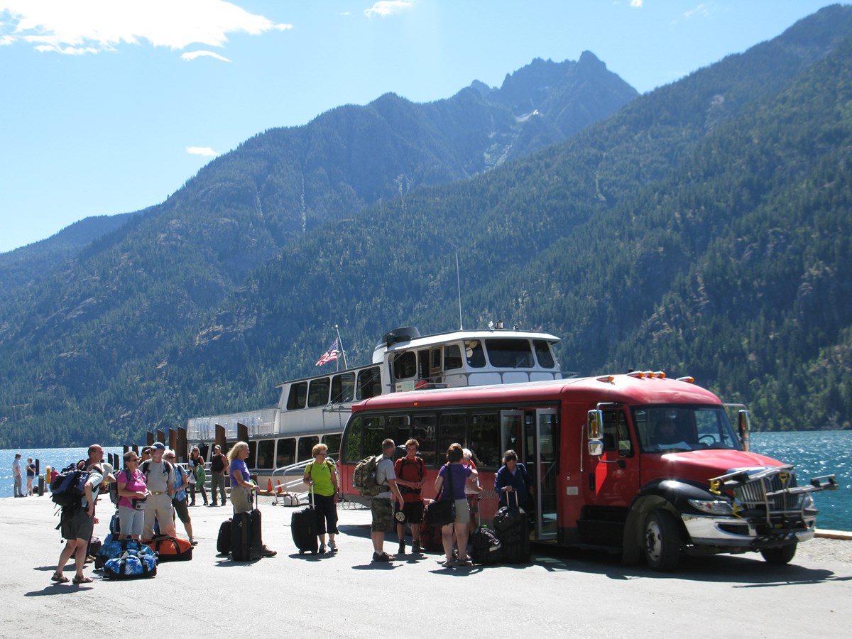 A group of people wait in line at a ferry dock to load a large red bus