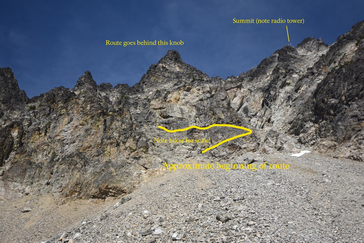 A multi-cragged mountain of gray rock rises from a talus field. A yellow line marks a possible route up.