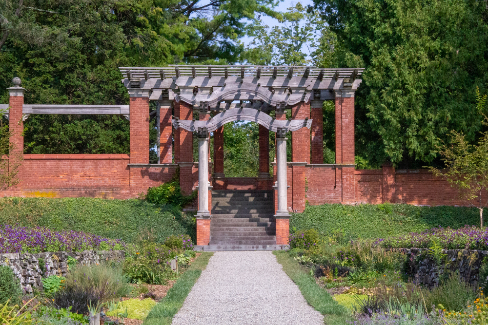 A brick pergola at the foot of a gravel path lined with garden beds.