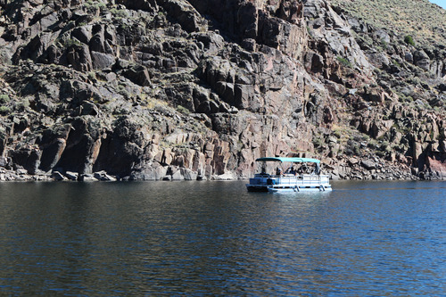 A white pontoon boat with a green awning crosses a flat section of water. Tall, rocky cliffs rise from the water's edge.