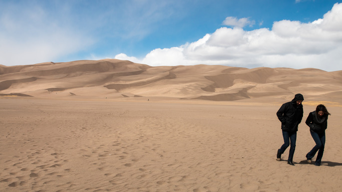 Two people walk across the flat sand field below the Great Sand Dunes. 
