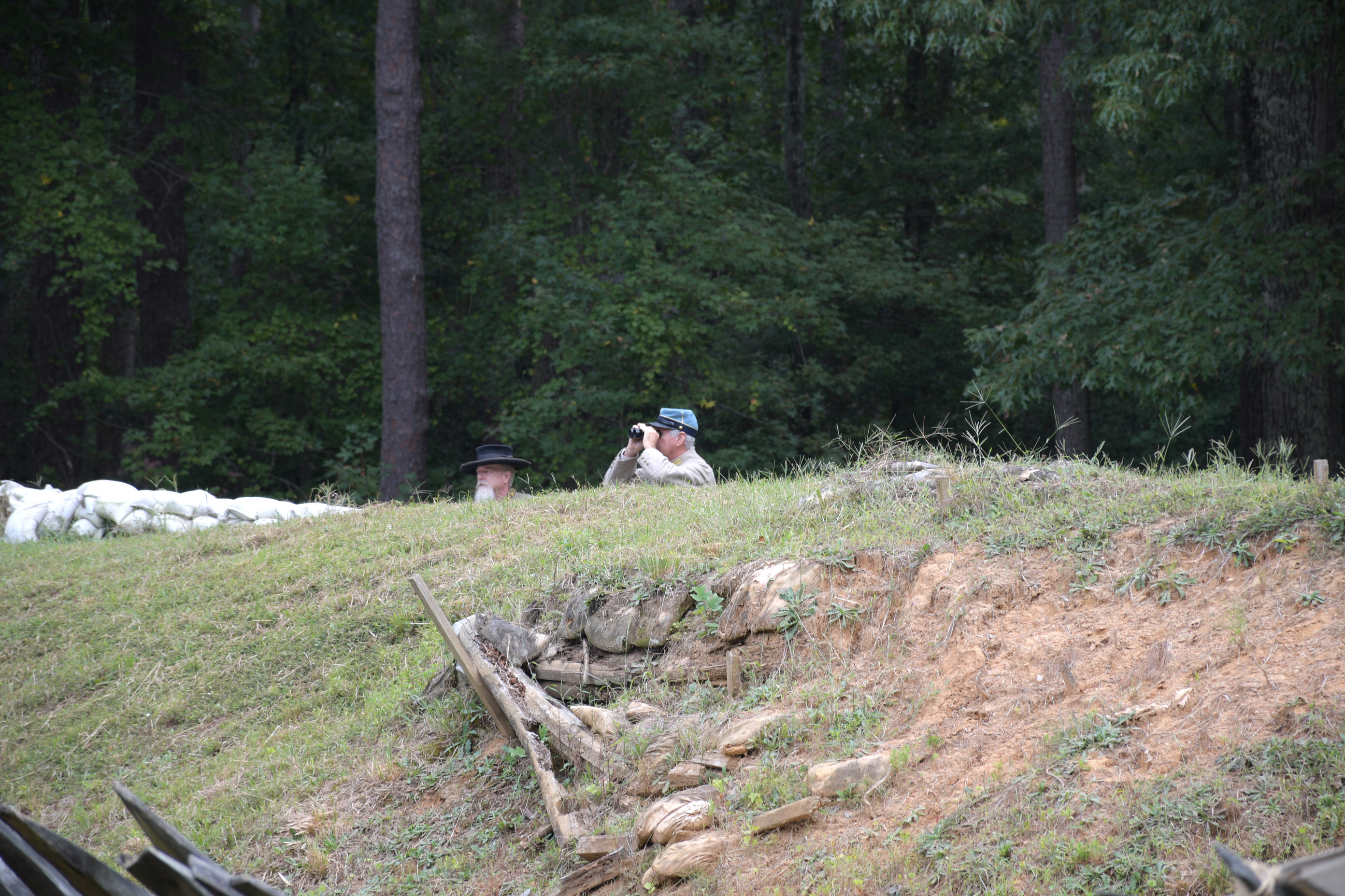 The heads of two white men wearing Confederate uniforms are visible above earthworks. One man is using a pair of binoculars.