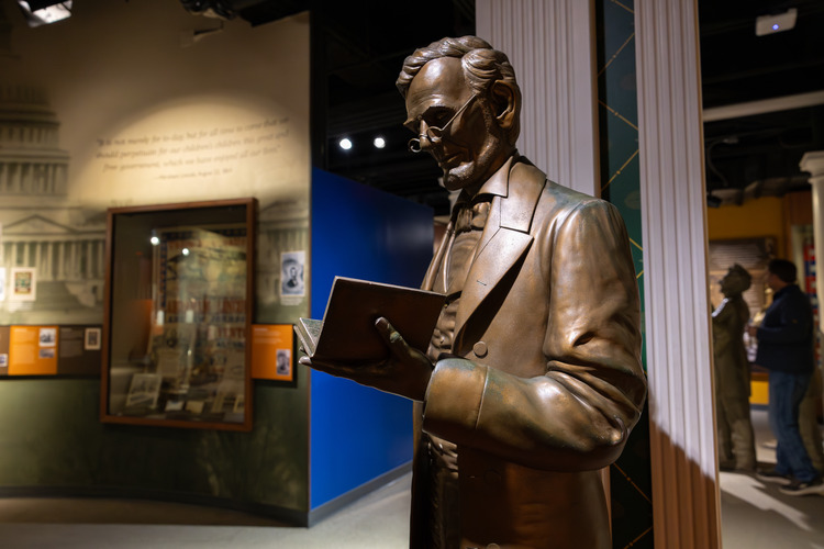 A bronze-colored statue of Lincoln wearing a suit and reading a book in his hand in a room with museum exhibits along the walls.