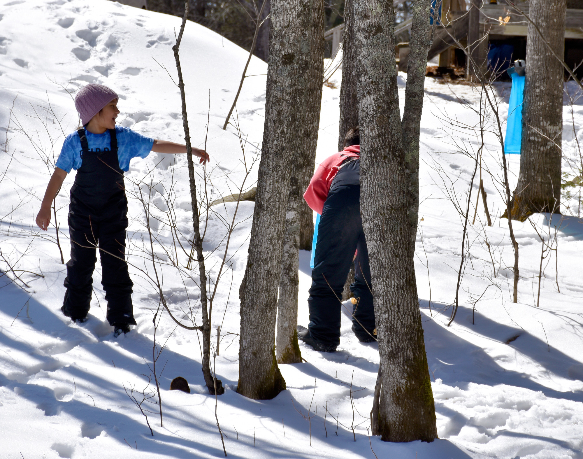 A child wearing snow pants and a knit hat points near another child in snow pants holding a blue plastic bag, leaning toward a tree.