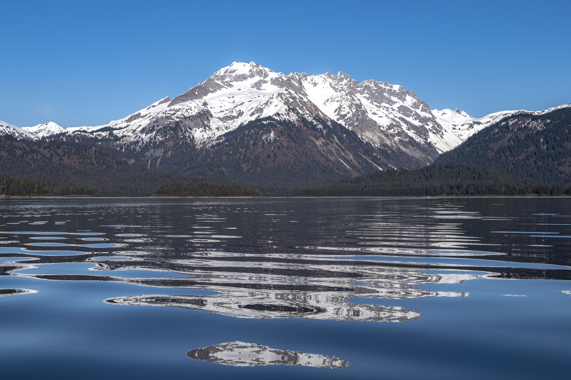Snow-covered mountains rise from calm water that reflects the scenery in abstract shapes.
