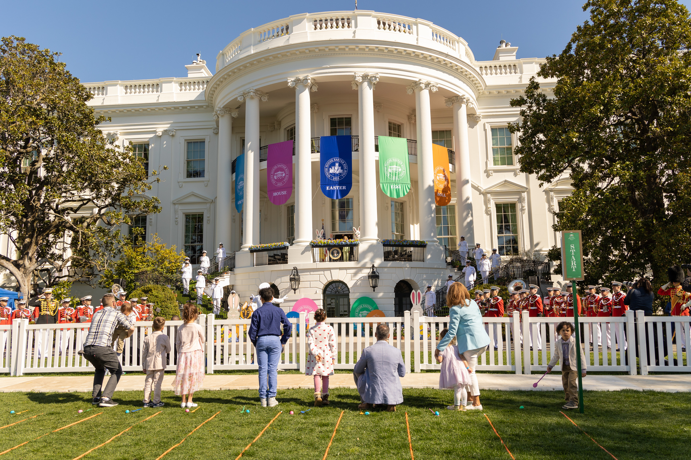 Visitors stand on the South Lawn of the White House watching the president and first lady give a speech. 