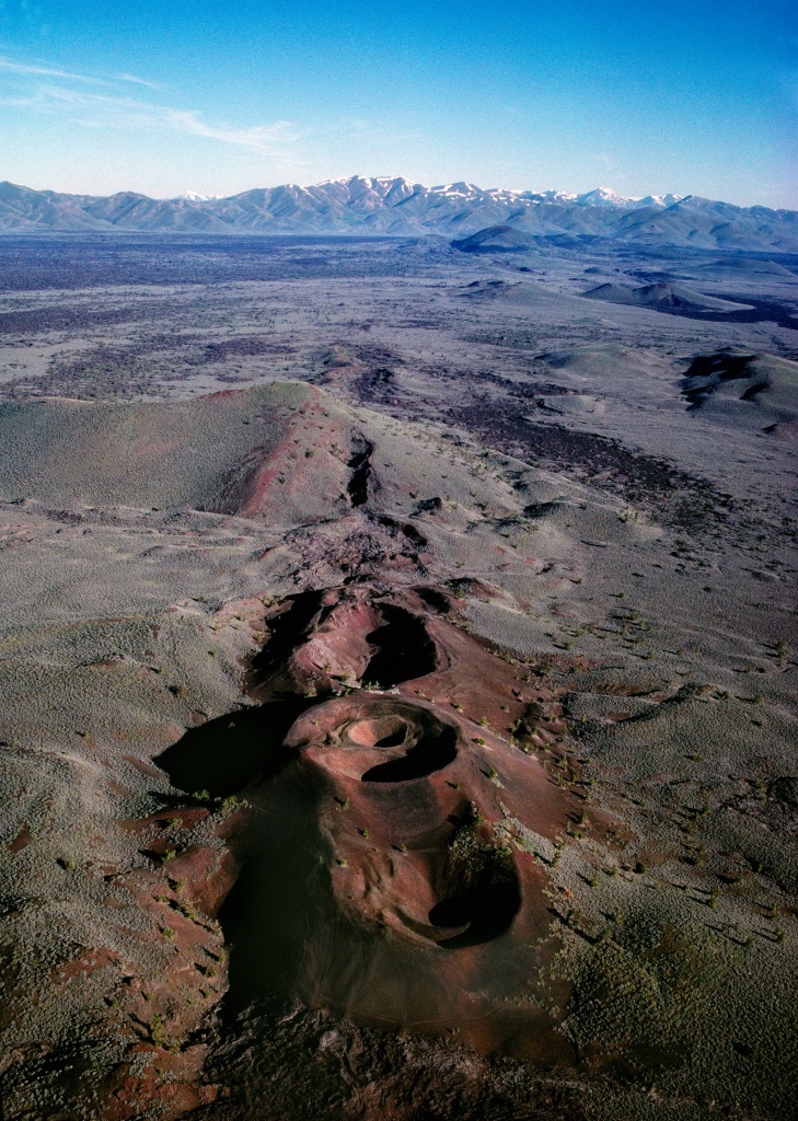 A chain of several round, volcanic craters along a ridge in the landscape leading toward a distant mountain range. One crater has another, smaller crater inside. Several other cinder cones are visible in the distance.