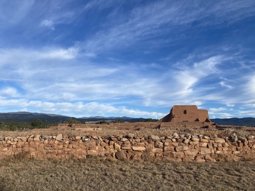 A stone wall stands between a patch of winter grass and the adobe remnants of the Spanish Mission and Convento. The Sangre De Cristo Mountains rise up far in the background under a blue sky.