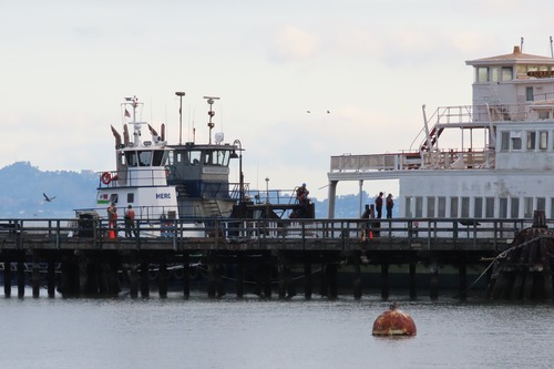 Alongside Hyde Street Pier, two tow vessels sit at Eureka's bow. Staff stand on the ferryboat and on the pier.