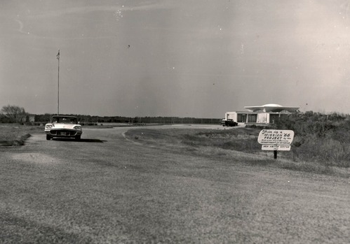 Black and white photo of roadway leading to visitor center. 1960s era car seen driving towards the viewer. Visitor Center is seen in the background at right with 1960s era truck parked in front. Sign in midground notes that the building is a Mission 66 project.