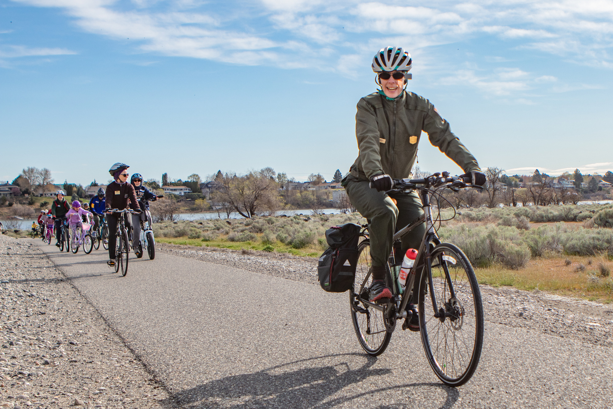A group riding bicycles on a paved path led by a ranger in uniform and helmet, framed by fields, blue skies, and a river in the background.