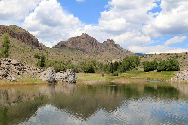 Mesas and pinnacles reflected in a body of water. Blue sky and clouds are above the scene.