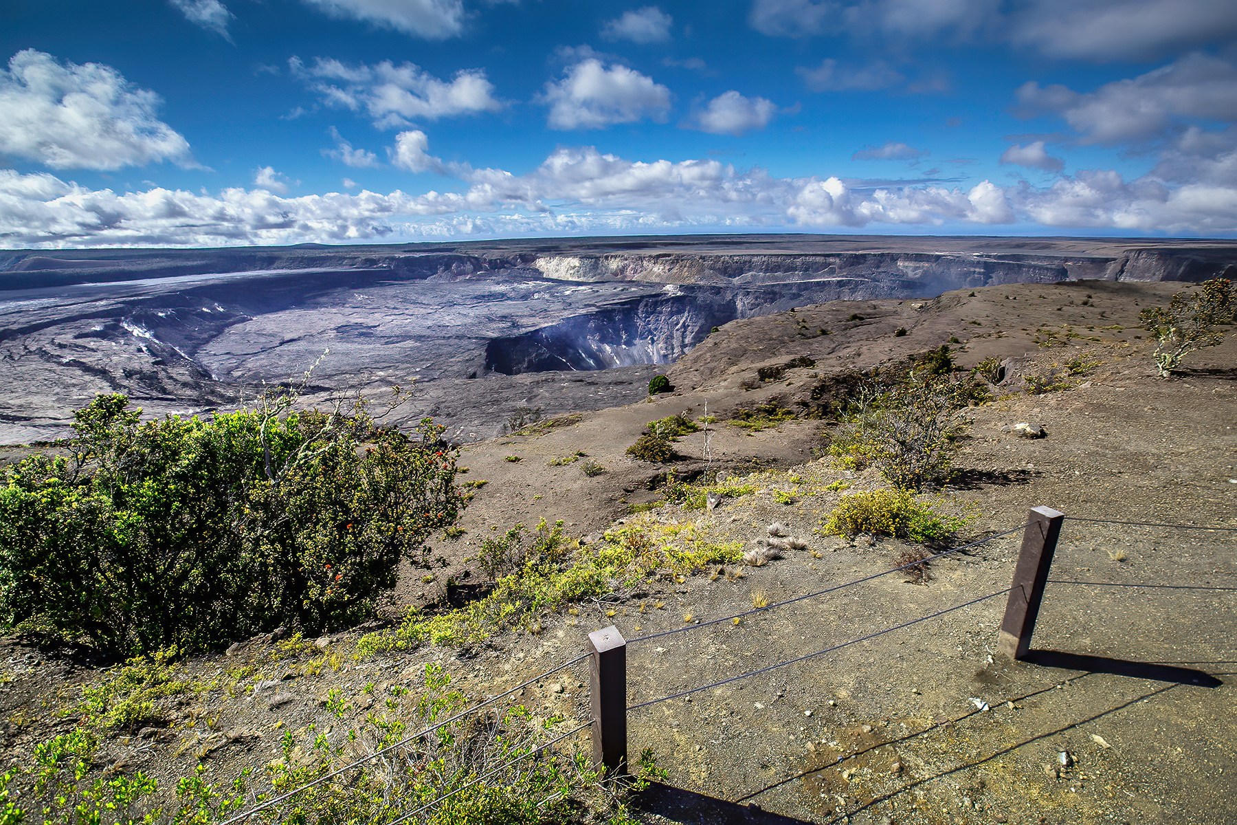 A non-erupting volcanic crater is seen from behind post and cable barriers at the edge of a cliff. 