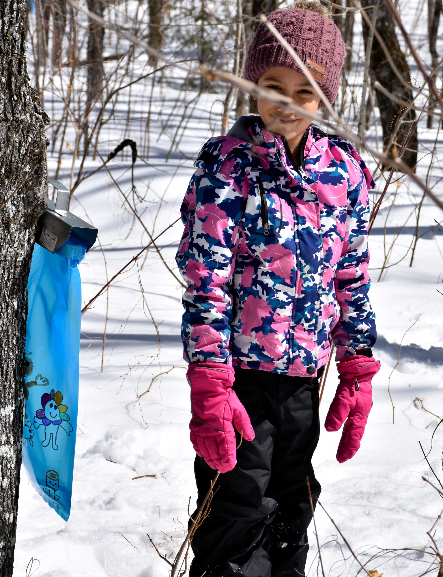A child wearing a pink and blue patterned jacket and mauve knitted hat smiles standing next to a blue plastic bag hanging from a tree.