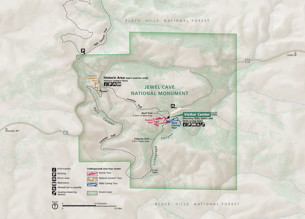 Map of Jewel Cave National Monument (NM) in South Dakota. The park is highlighted and outlined in green, and the surrounding terrain is tan speckled with green to represent forest cover. Black dashed lines represent trails, and white lines represent highways and roads.