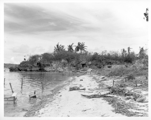 A low rocky point stretching into the ocean. In the base is a large man-made hole that was used as a pillbox. In the foreground, the water and the beach are lined with debris and barbed wire.
