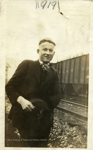 A portrait of a man, the image is cut off at his legs. He wears a suit next to train tracks witch a box car behind him. He is smiling slightly and is holding a wide-brimmed hat flat against his torso.