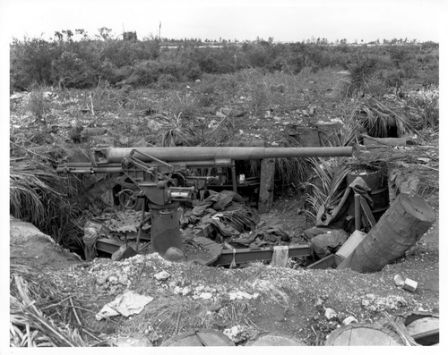 A large gun with a gunner seat sits in a lower bunker made of logs and sandbags. Behind the gun is debris from bombing.