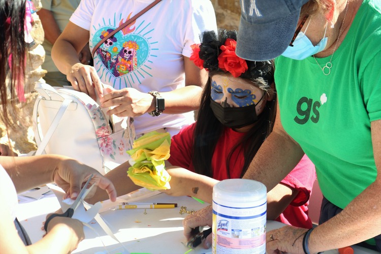Girl with her face painted like a sugar skull making a paper marigold flower at a table. 