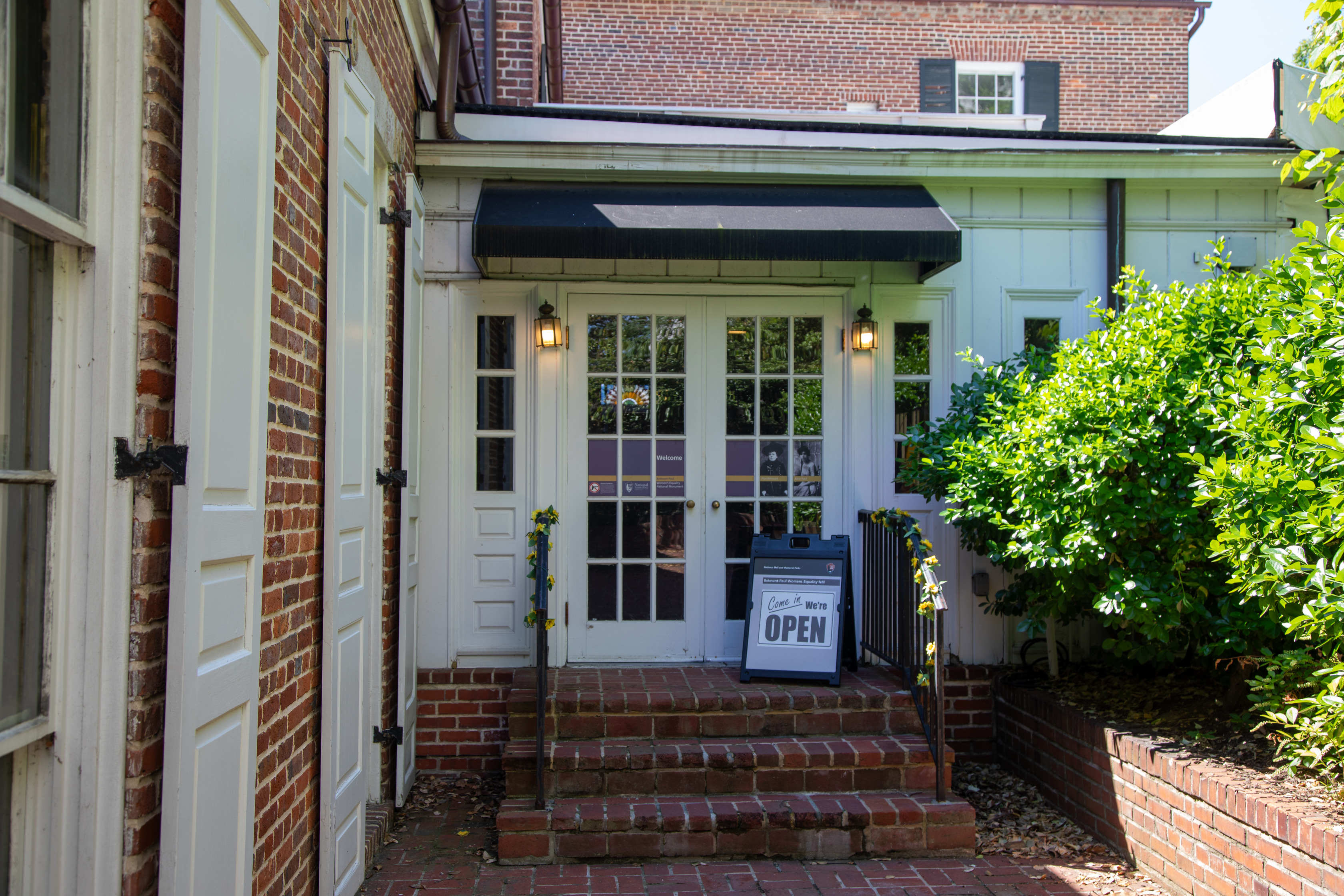 Three brick steps with handrails on each side lead up to a door with windows and round knobs on the side of a brick building.