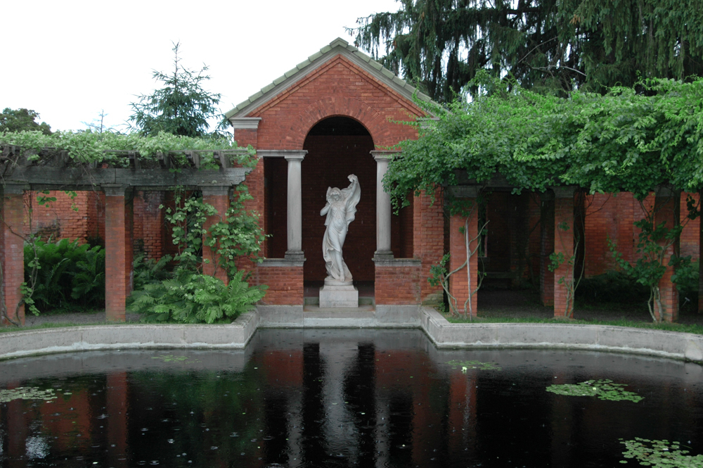A reflecting pool in front of a brick pergola with marble statue of an woman in dance pose.