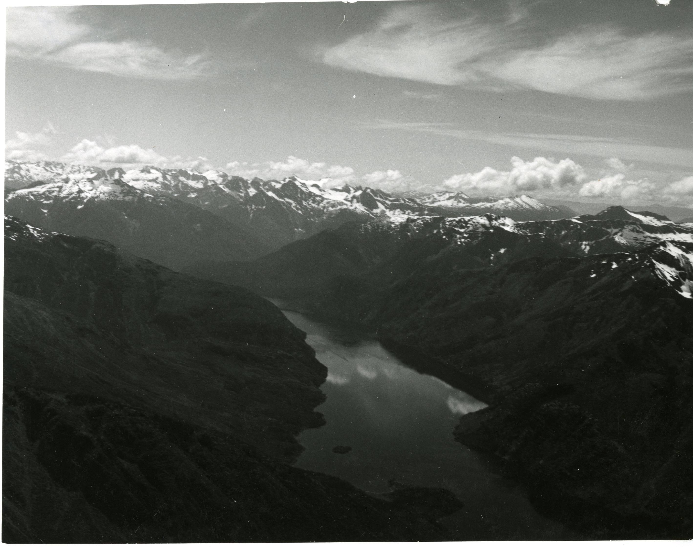 Aerial view of a mountain range, with a lake in between two mountains. Mountain peaks covered in snow and ice stretch into the horizon.