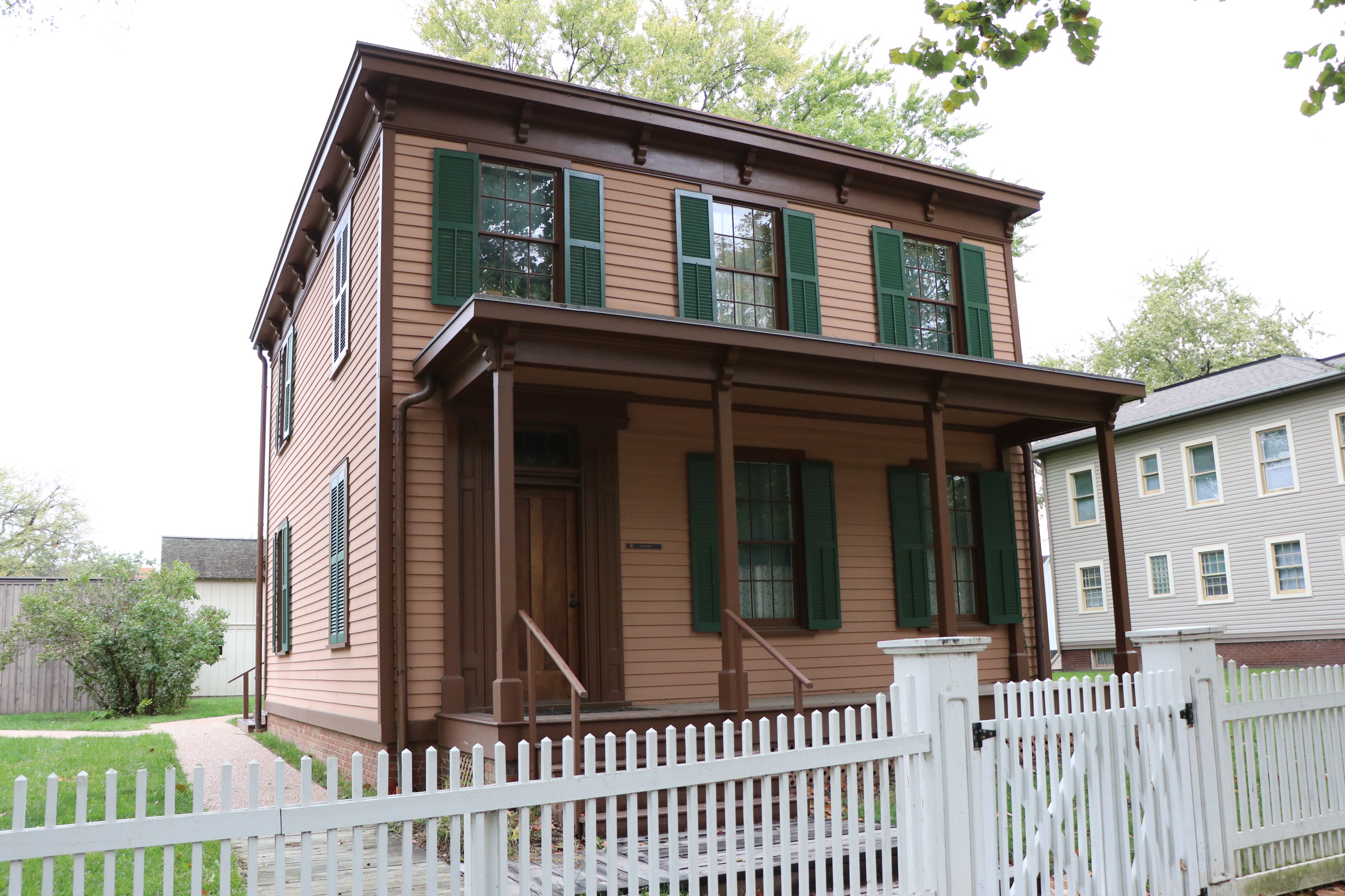 A two-story light brown house with green shutters and a white fence in front of it.