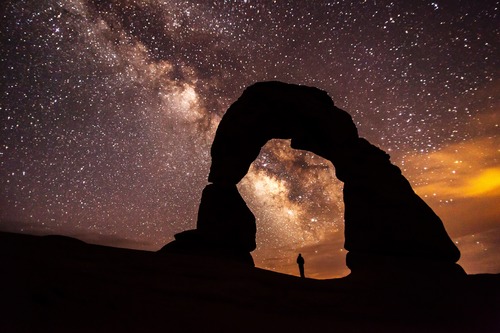 the milky way and a starry sky above a silhouetted stone arch and a person standing beneath it