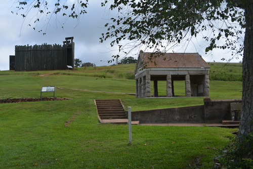 A stone structure sits on a hill in front of a wooden stockade.