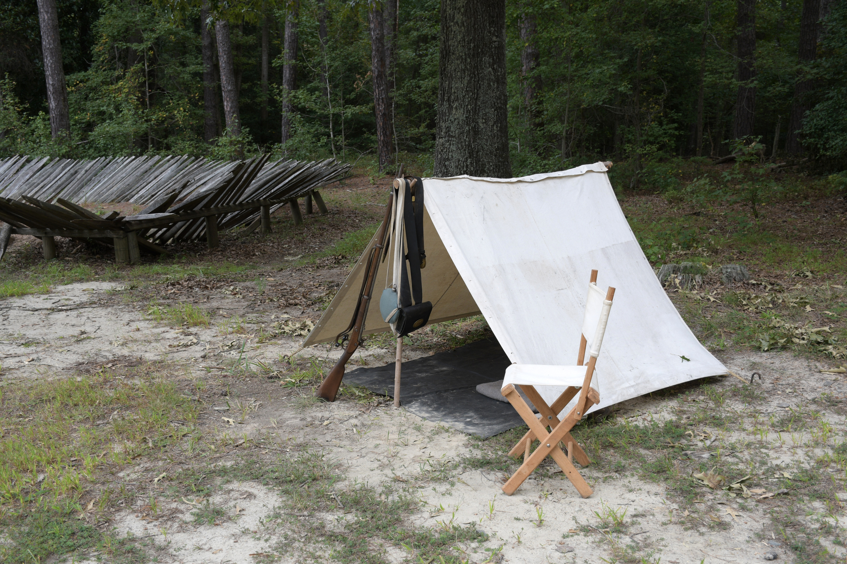 A rifle leans against an upright support pole of a tent. A canteen and cartridge box hang from the same support post. A white canvas folding chair stands outside the front. A wooden palisade is in the right background. 