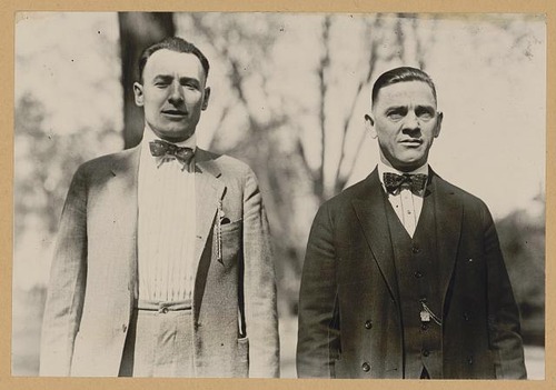Two men stand next to each other with their arms aby their sides. They are wearing early 20th century suits, the man on the left in a gray suit and the man on the right in black. The man on the left is taller and is smiling, whereas the man on the left has a serios expression and is five to six inches shorter. The photo was taken outdoors.