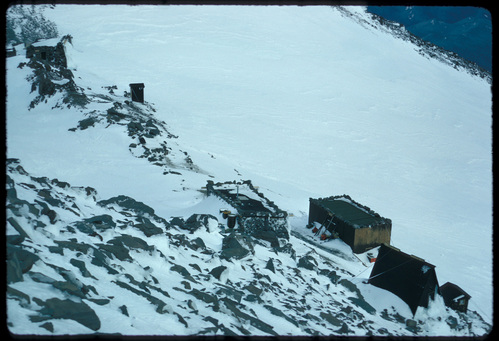 A collection of small buildings on a rocky ridge, some built out of snow and others of wood, all partially covered by windswept snow.