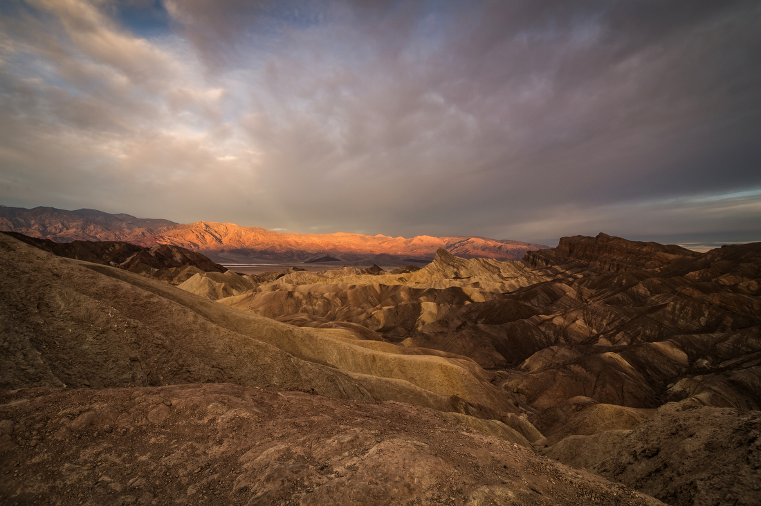Striped yellow and brown hills of bare stone, with sunlight beginning to hit distant mountains and a mostly cloudy sky.