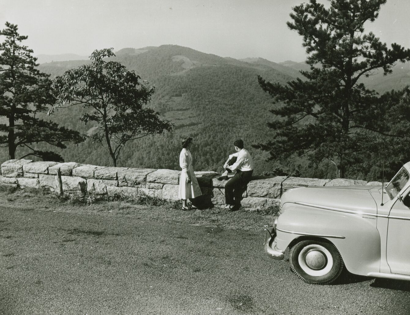 Two people sit on a stone railing looking out along a mountain ridge