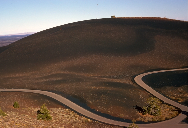 a road winds alongside a smooth black cinder cone with a few small plants along the road, two small people can be seen walking down one side