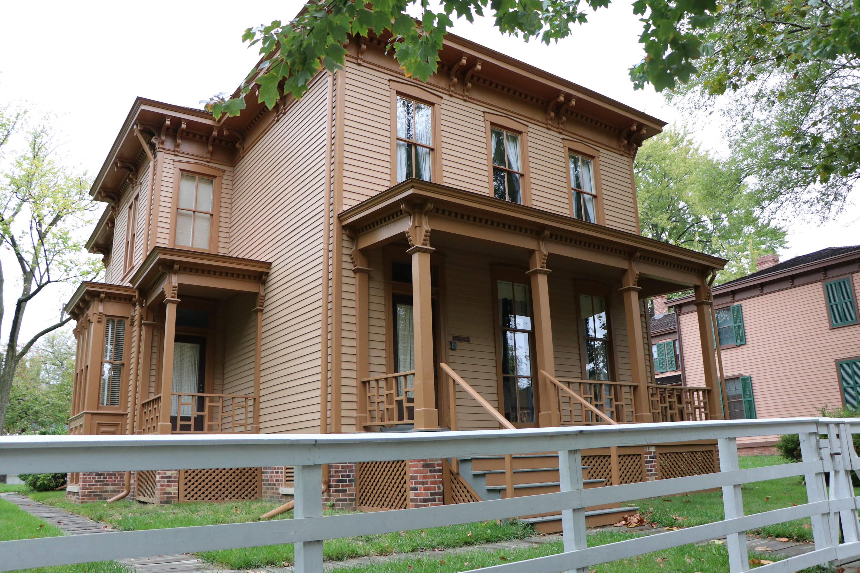 A two-story tan house with large windows and a white fence in front of it.