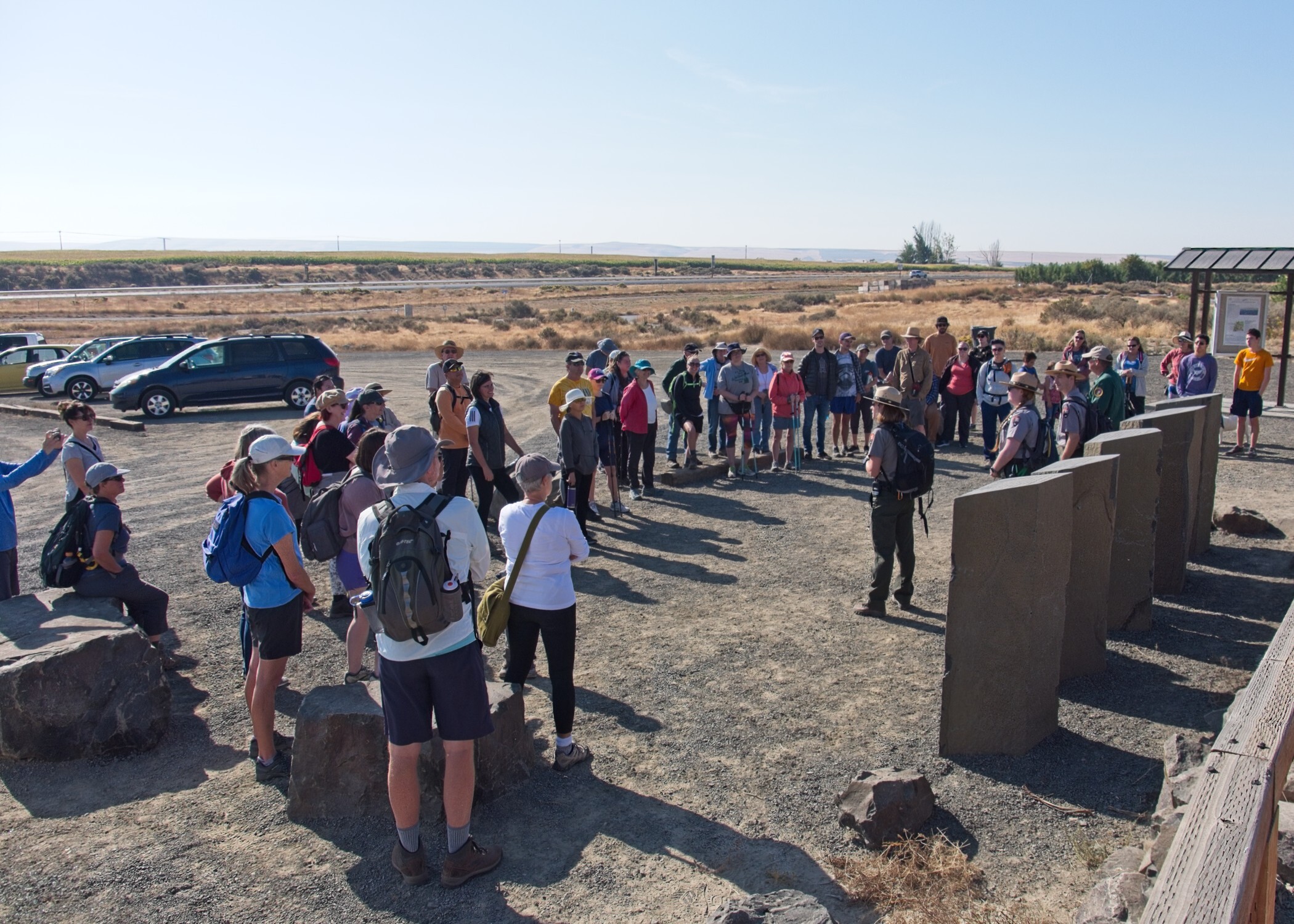 Three uniformed rangers stand surrounded by about fifty participants in a gravel lot trailhead with dried grass fields extending to the horizon.