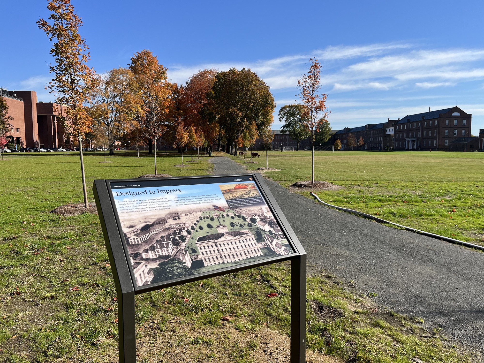Wayside titled "Designed to Impress" overlooking the parade grounds of the Armory. Several brick buildings are visible in the distance beyond the stretch of green grass, asphalt path and cluster of trees in shades of red and brown. 