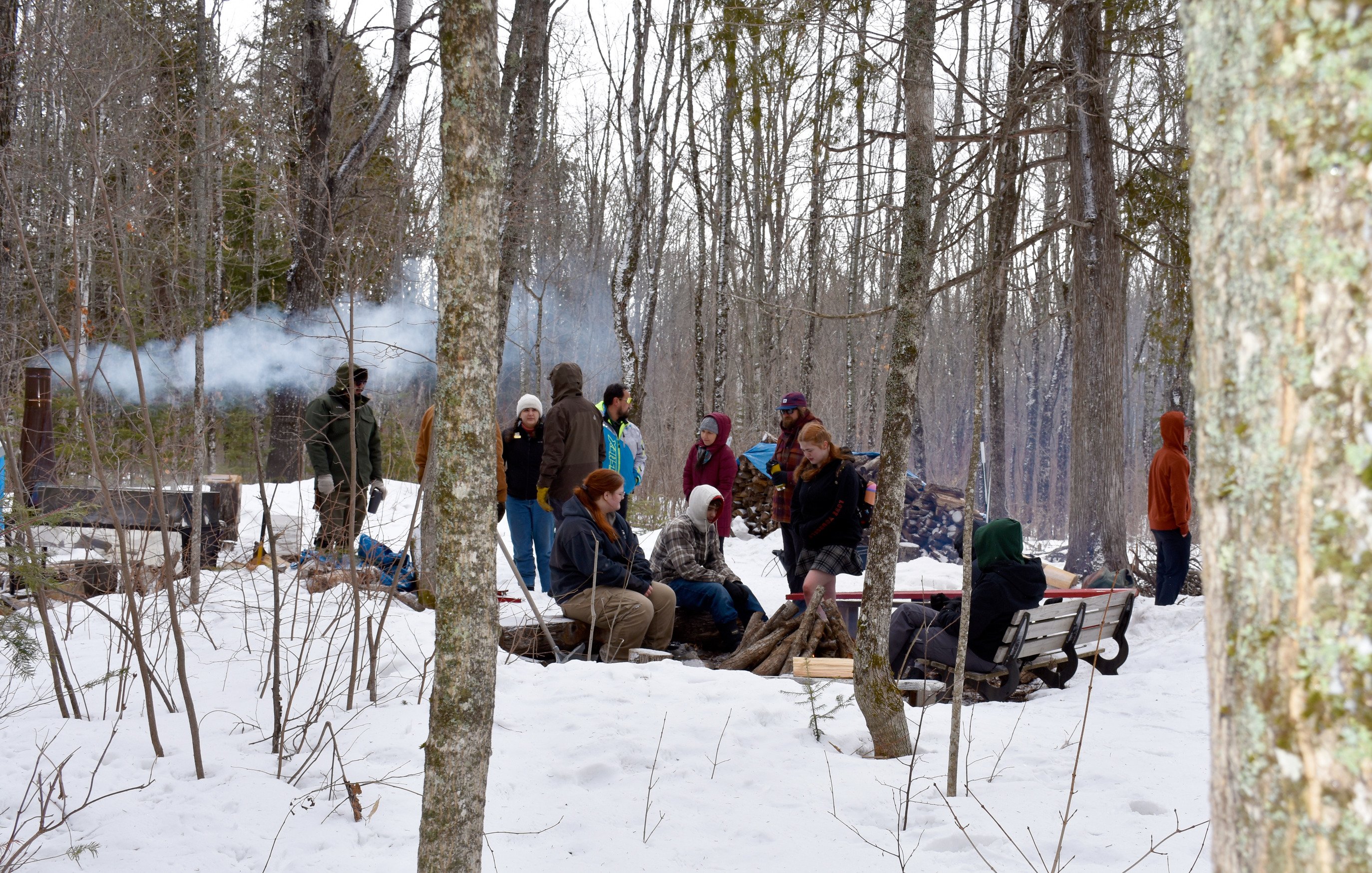 Several people in winter clothing sit and stand near a fire next to a steaming metal unit, surrounded by leafless trees.