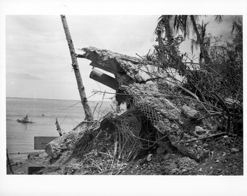 The barrel of a large gun pokes out of hole camouflaged with pieces of concrete and wire. The ocean is in the background.