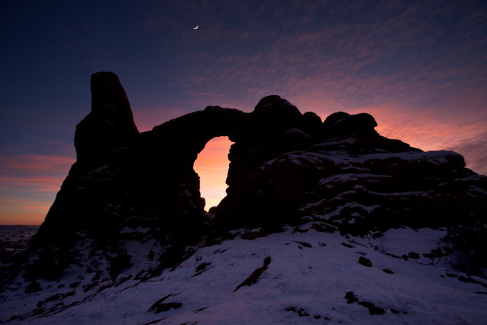 a crescent moon rises above a silhouetted stone arch with snow on the ground