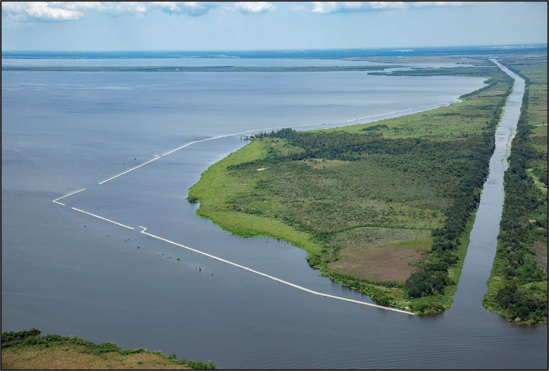 An aerial view of Jean Lafitte National Historic Park and Preserve Shoreline featuring green earth surrounded by a water - a canal on the left side and open water on the right and foreground.